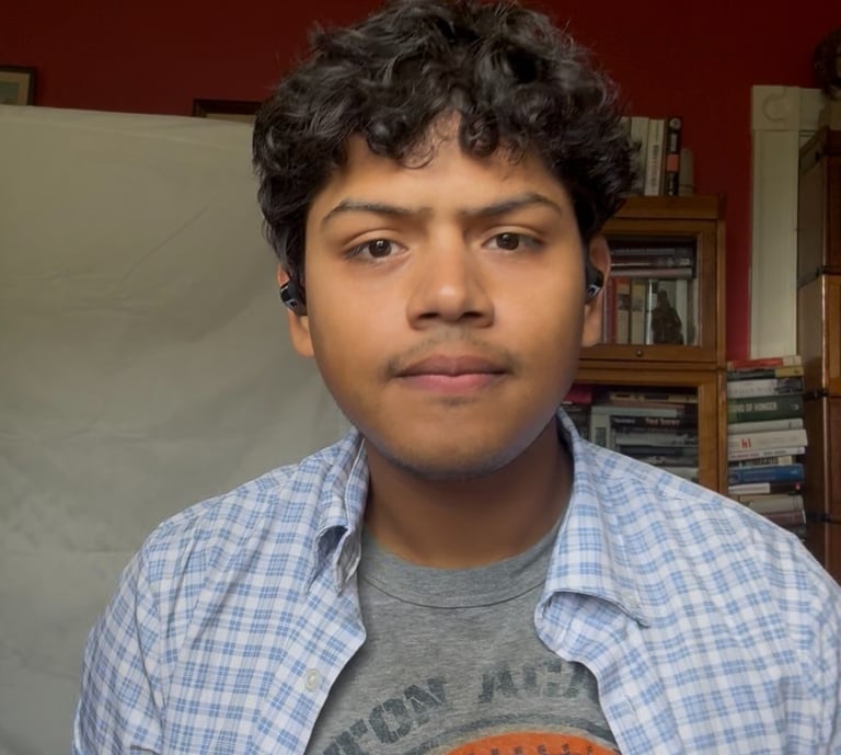 Young man with curly hair wearing a blue plaid shirt and wireless earbuds in a room.