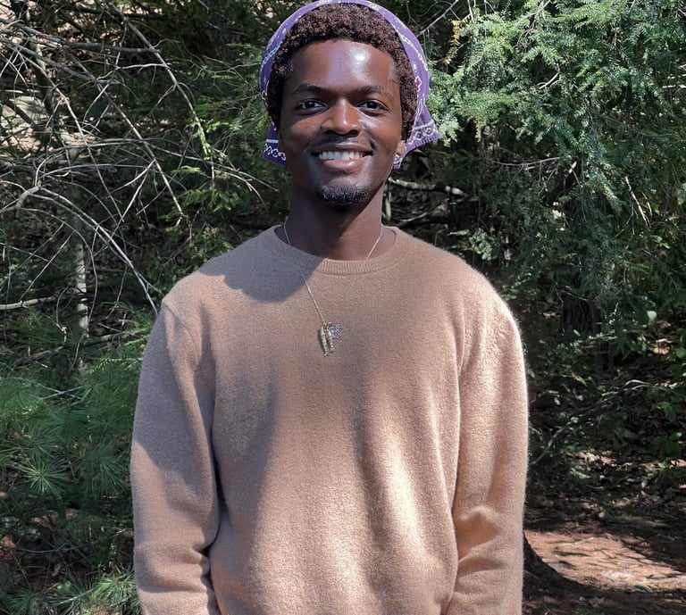 Smiling young Black man wearing a tan sweater and purple bandana in a forest setting.