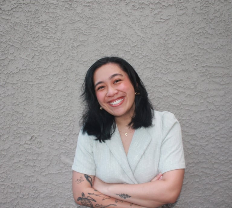 Smiling woman with black hair and arm tattoos posing against a textured grey wall.