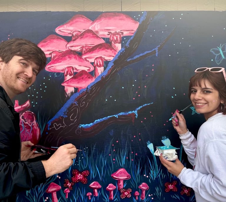 Two artists painting a whimsical mural featuring glowing pink mushrooms and butterflies.