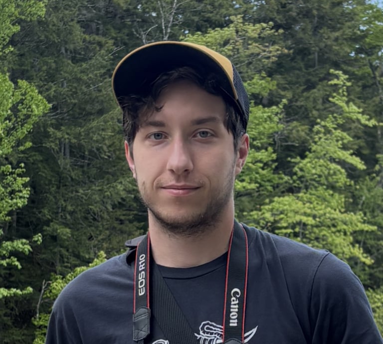 Young male photographer wearing a yellow hat and Canon EOS R10 camera outdoors with forest background.