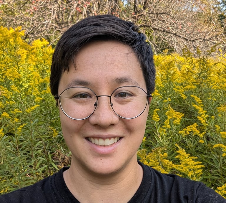 Smiling transgender man with short hair and glasses in front of blooming yellow wildflowers.