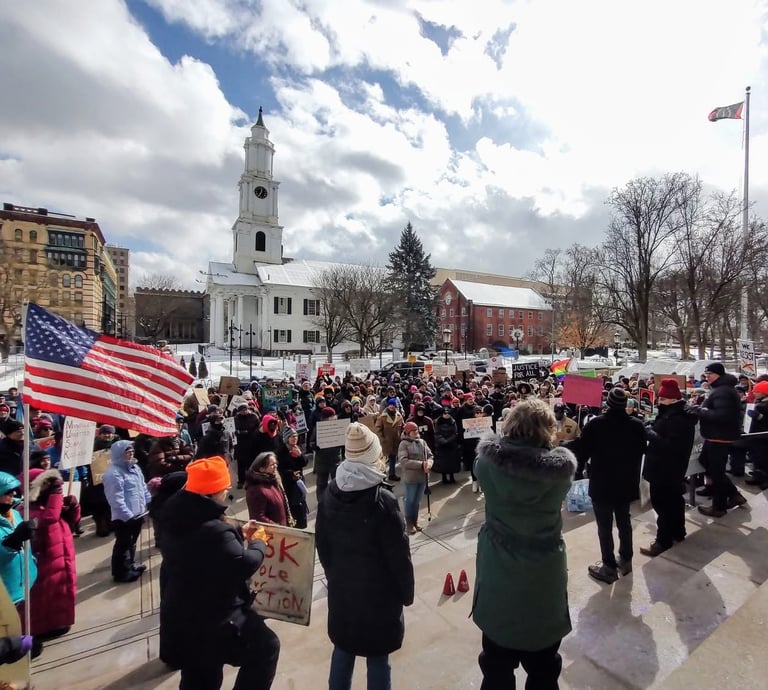 Group of people rallying on a cold snowy day. A church in the background. An American flag flying. 