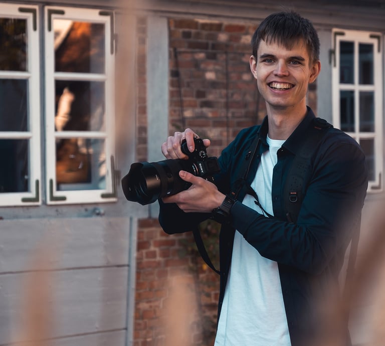 Lächelnder Hochzeitsfotograf Joel Pingel mit Kamera vor Backsteingebäude. Portrait: Just Perspectives.