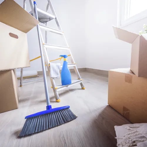 A ladder with a spray bottle and cleaning rag, a broom and brown moving boxes stacked in an empty room.