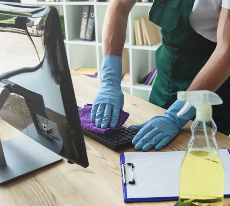 Man wiping down keyboard while wearing cleaning gloves, green apron. Office setting in the background with cleaning supplies
