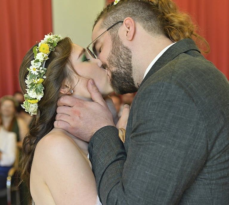 Ein Brautpaar küsst sich während der Trauung innig vor den applaudierenden Hochzeitsgästen. Die Braut trägt ein Blumenkranz i