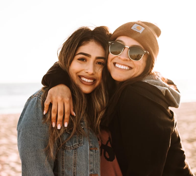 Two young women smiling and embracing on a beach at sunset, one wearing a denim jacket and the other in a beanie & sunglasses