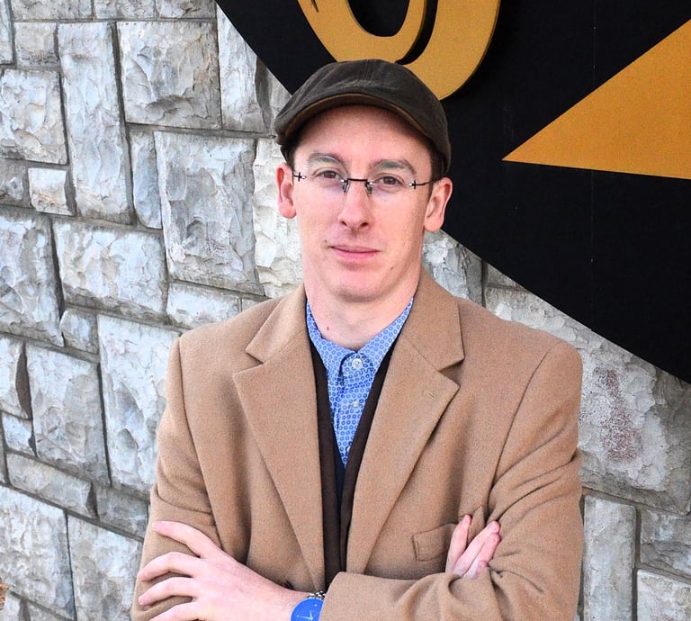 Professional man wearing a tan overcoat, flat cap, and glasses posing against a stone wall.