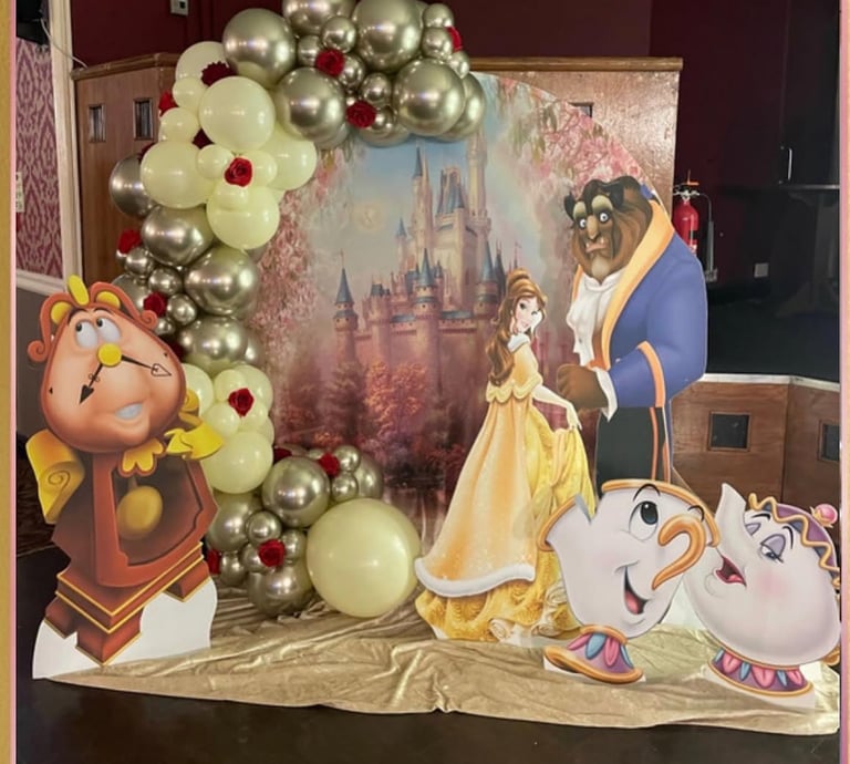 Beauty and the Beast party decorations featuring a gold balloon arch and character cardboard cutouts.