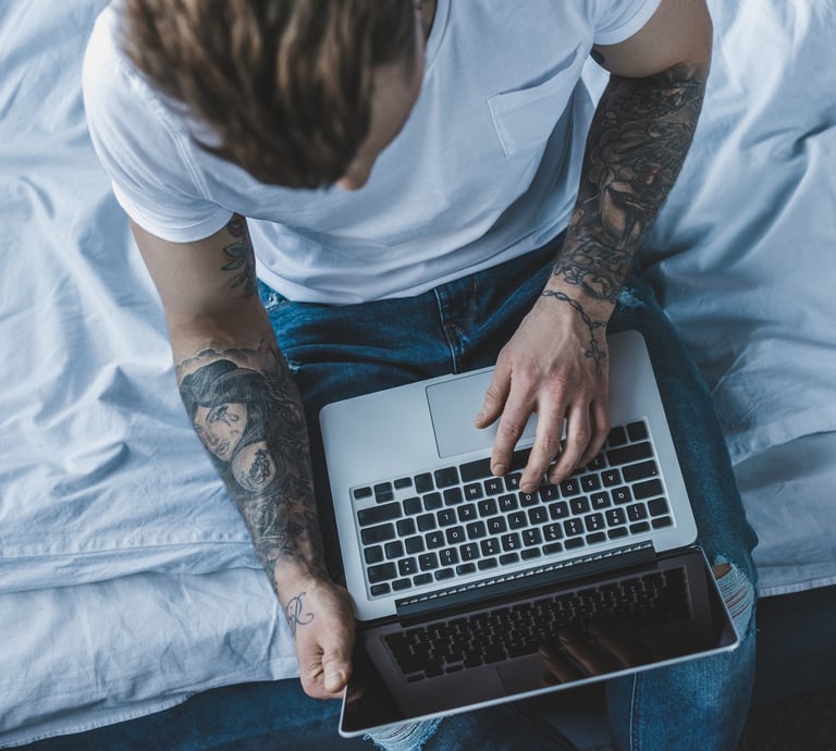 man with tattoos on arms holding a laptop while sitting on a bed