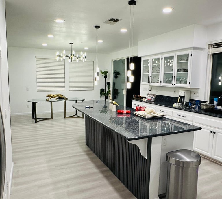 A modern kitchen with white cabinets, a large island, and a dining table, decorated with vases, book