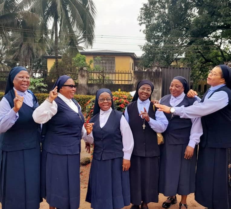 A group of cheerful Catholic nuns in blue and white habits smiling and socializing outdoors.