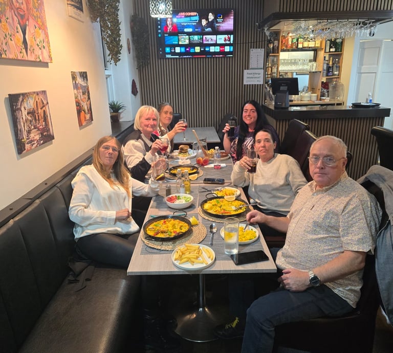 A group of smiling customers enjoying paella and drinks together in a cozy Spanish restaurant.