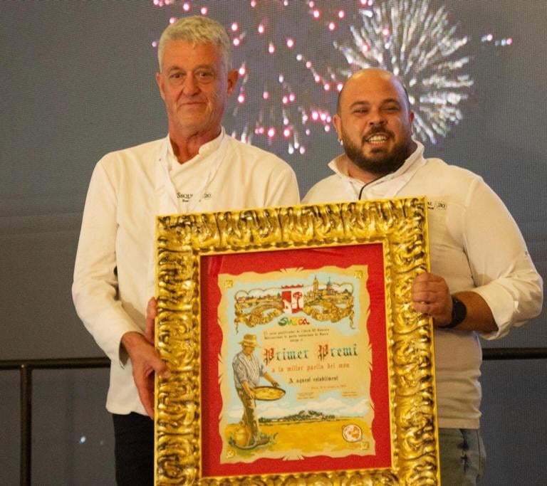 Two chefs proudly holding a gold-framed first prize award certificate for the best paella in the world.