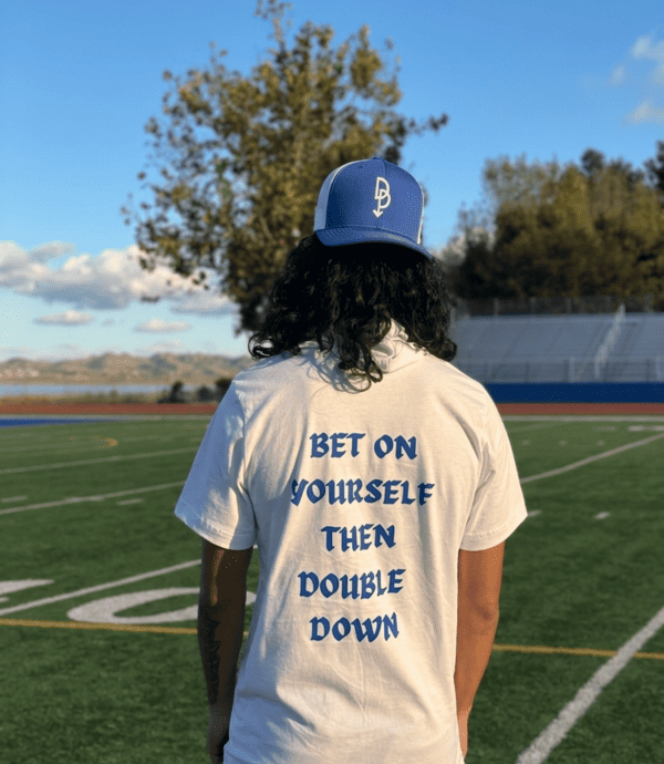 A person wearing a Bet On Yourself graphic t-shirt and blue trucker hat on a football field.