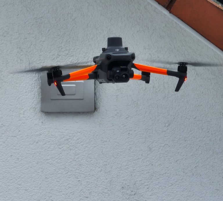 A DJI Mavic drone with orange arms flies near a building wall for an aerial roof inspection.
