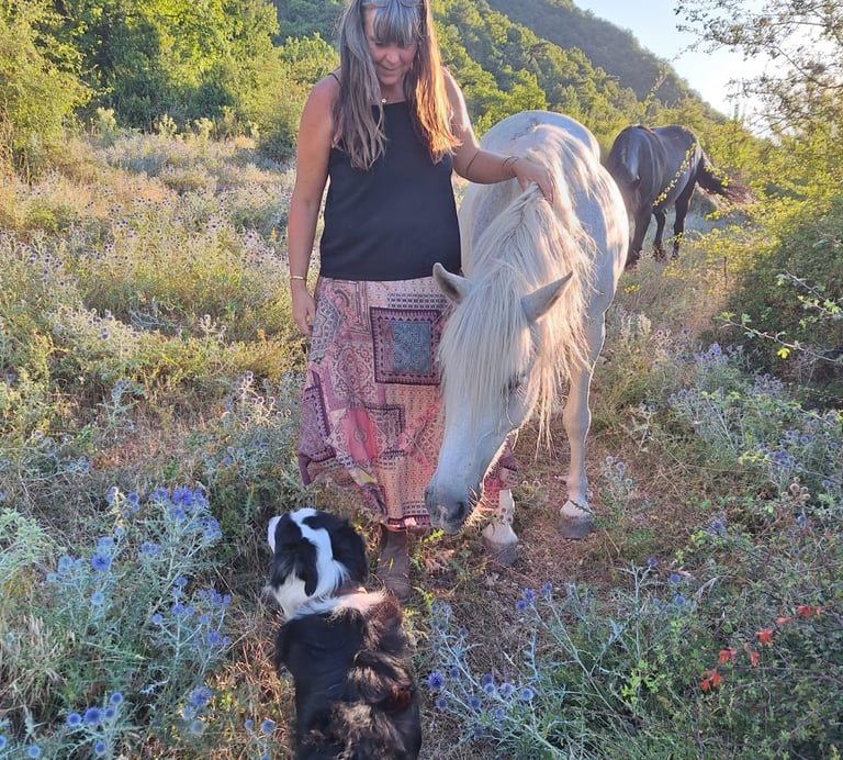 Vrouw staat naast een wit paard in een weiland met bloemen, ze legt haar hand op het paard