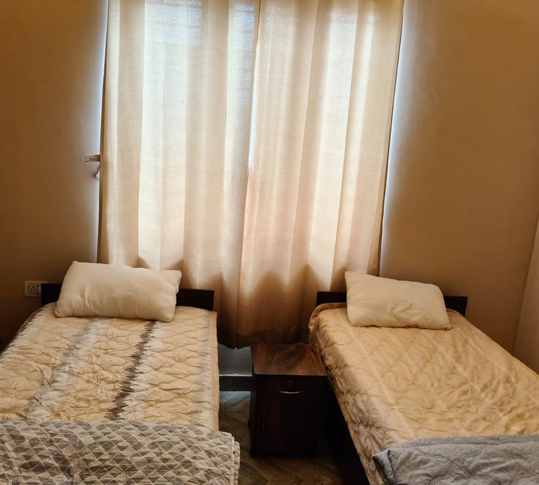 Two single beds with beige patterned bedding and white pillows in a cozy bedroom with light brown curtains.