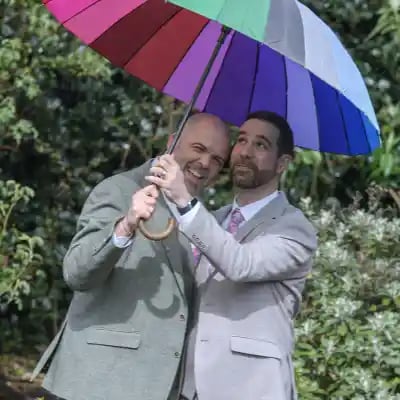 A happy groom couple in suits posing together under a colorful rainbow umbrella at their wedding.