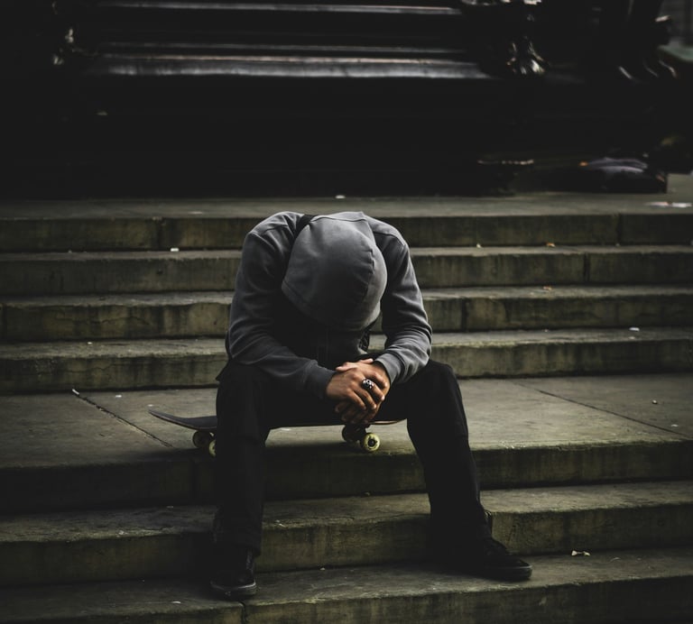 A lonely skateboarder in a grey hoodie sits on his board on city concrete stairs in a moody urban setting.