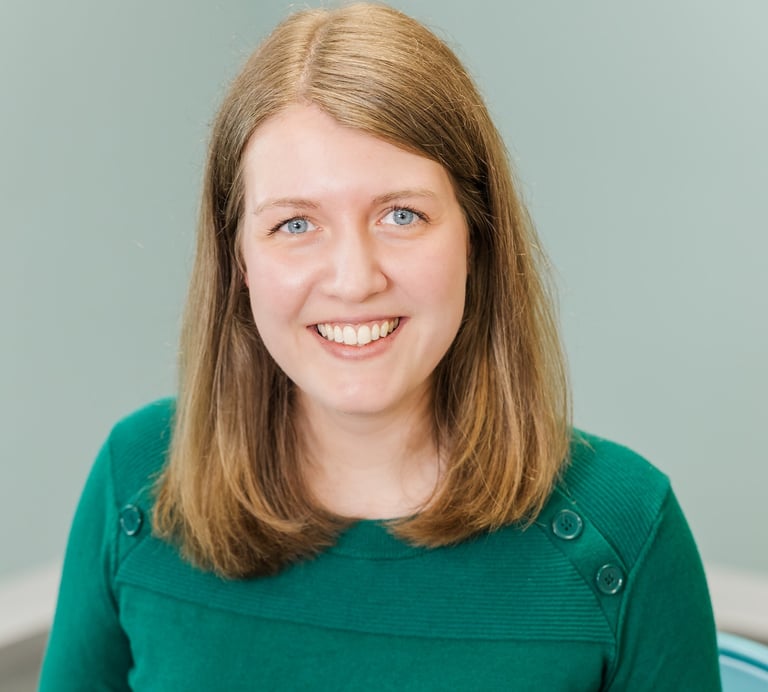 a woman in a green shirt and a blue chair. She has dark blond hair and blue eyes and is smiling.