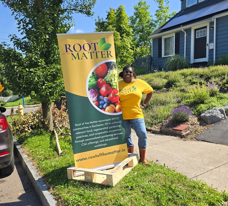 A woman in a yellow shirt stands next to a Root of the Matter banner for urban gardening in Rochester, NY.