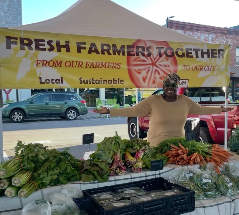 A smiling vendor at a local farmers market stands behind a stall with fresh sustainable vegetables.