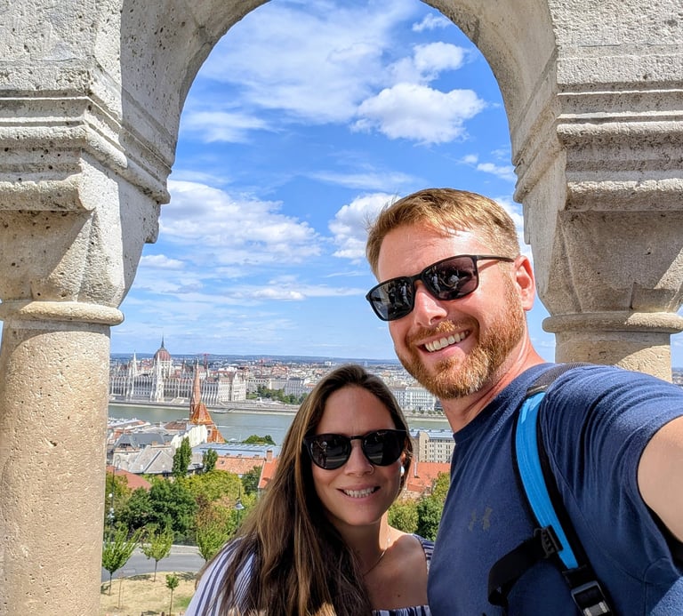 A smiling couple takes a selfie at Fisherman's Bastion with the Hungarian Parliament Building in the background.