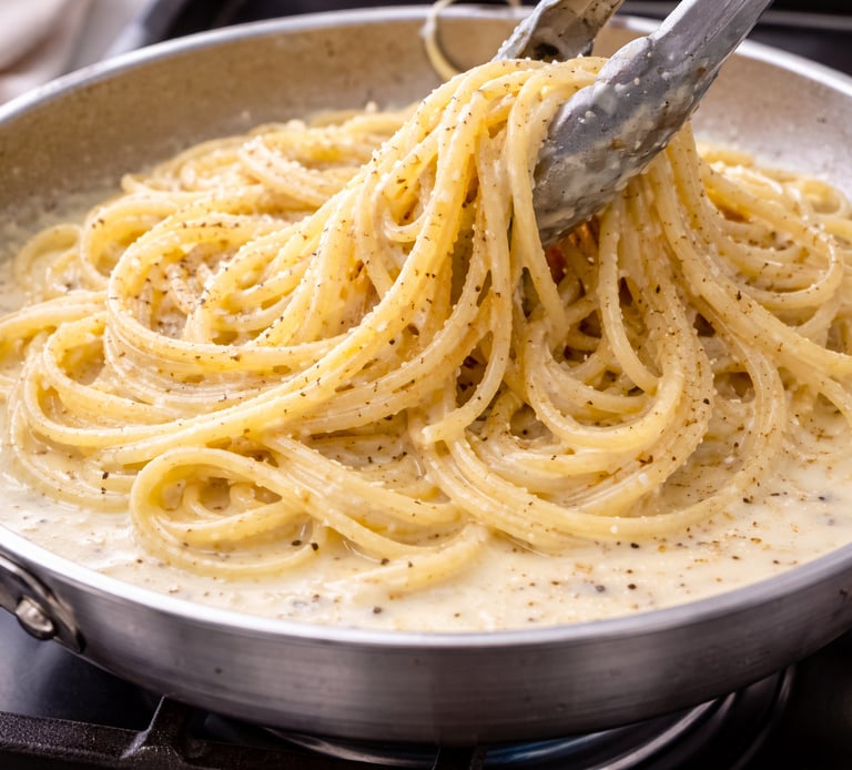 Tongs lifting creamy spaghetti cacio e pepe with cracked black pepper from a stainless steel skillet.