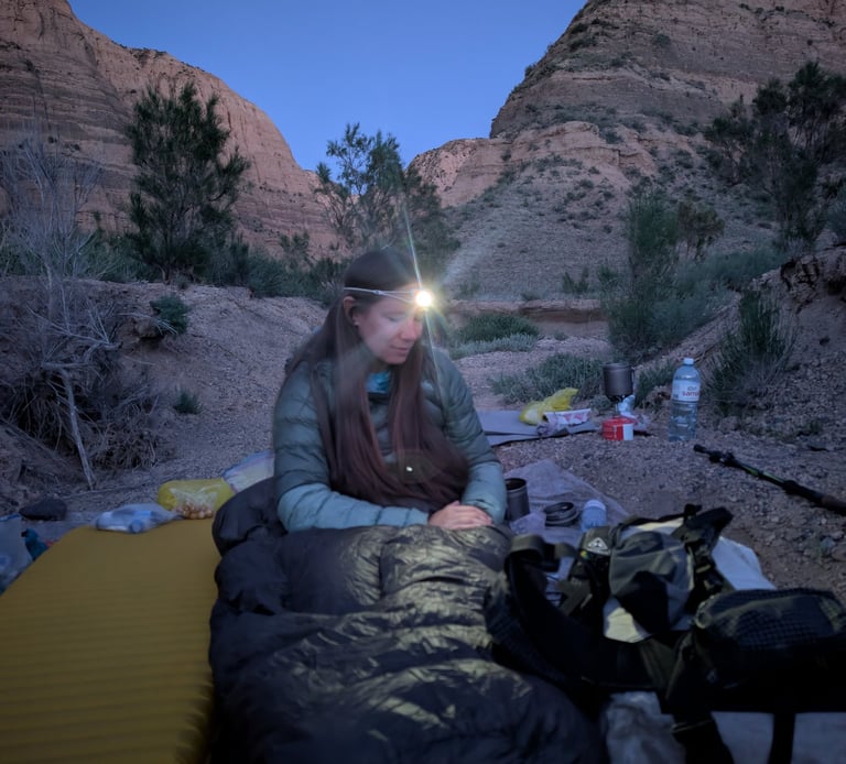 Bugs in her sleeping bag using her Petzl Bindi headlamp as dusk in a desert canyon campsite.