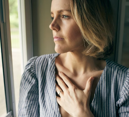 A woman gazes out a window with her hand on her chest, suggesting reflection and longing.