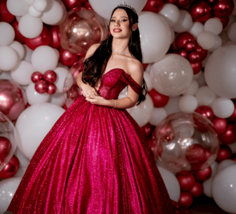 Debutante em vestido vermelho com tiara, em frente a painel de balões em cores vinho e branco, representando uma Festa de 15 
