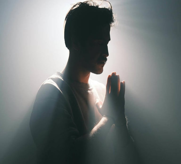 Silhouetted figure with hands in prayer, surrounded by radiant light against a dark background.
