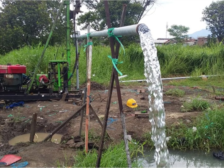 A portable water well drilling rig pumping a steady stream of clear water from a pipe into a muddy field.
