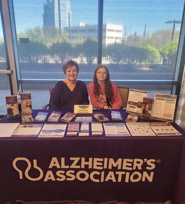 Photo of Debbie & colleague seated at an Alzheimer's Association info table, in front of a shaded window and urban landscape.