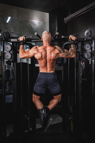 Muscular man performing wide grip pull-ups on a cable machine at the gym to build back muscles.