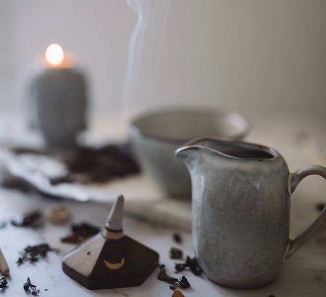 A ceramic incense burner releasing smoke next to a tea set and a lit candle for meditation.