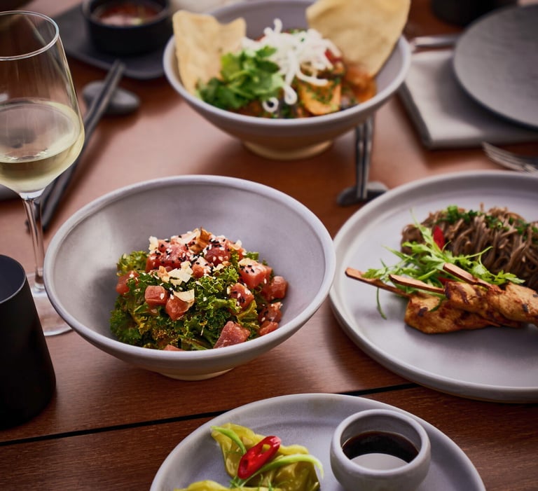 A gourmet spread of Asian fusion dishes including tuna tartare, chicken satay, and noodles served with white wine.