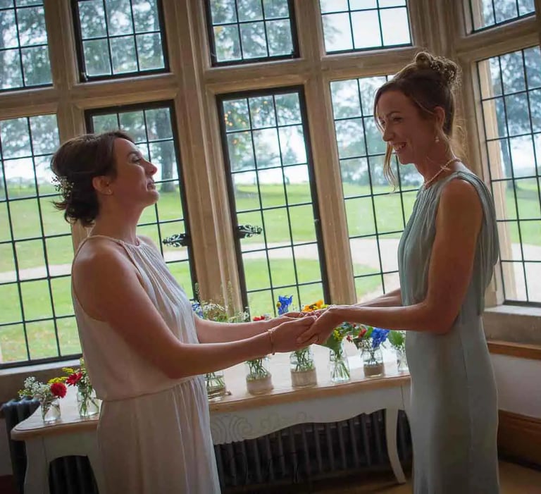 A happy same-sex couple exchanges vows during an indoor wedding ceremony by a large stone window.