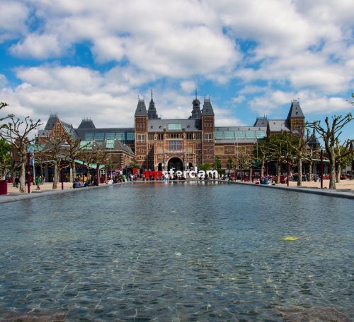The Rijksmuseum building in Amsterdam behind the Museumplein reflection pool under a blue sky.