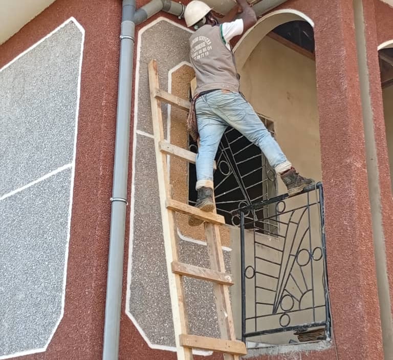 A construction worker on a wooden ladder installs a gutter drainage system on a building.