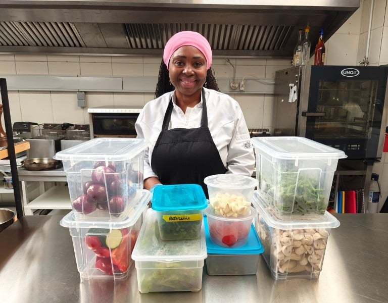 Chef Viviane smiling behind multiple labelled transparent prep containers filled with vegetables on a stainless steel.