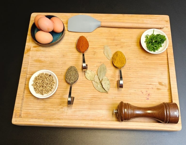 Overhead view of spices, eggs, fresh herbs and a pepper mill arranged on a bamboo chopping board on a dark