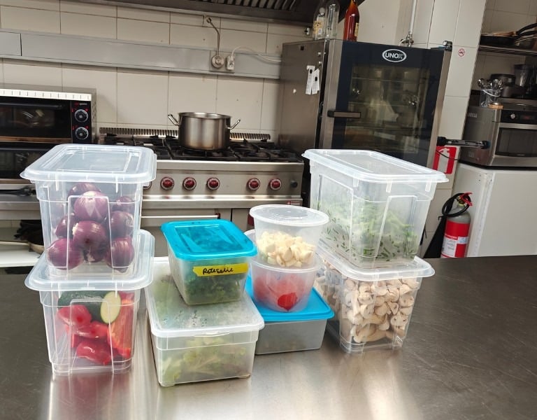 Multiple labelled transparent prep containers filled with vegetables and ingredients lined up on a stainless steel kitchen.