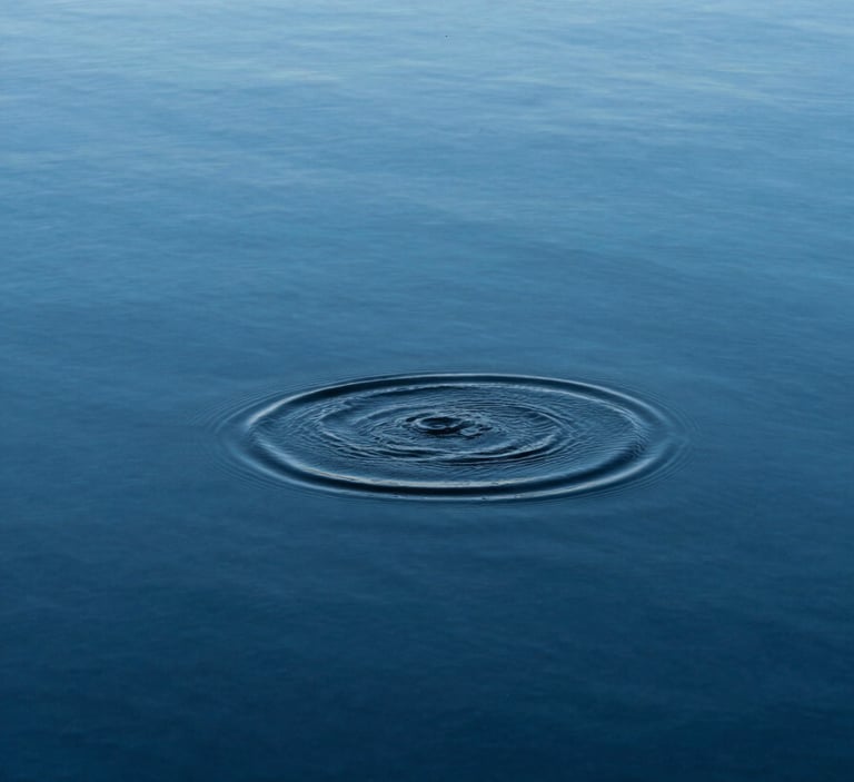 A minimalist, high-angle photograph of a single ripple moving across a perfectly still, deep blue lake. The water is crystalline, reflecting a pale azure sky and absolute stillness.