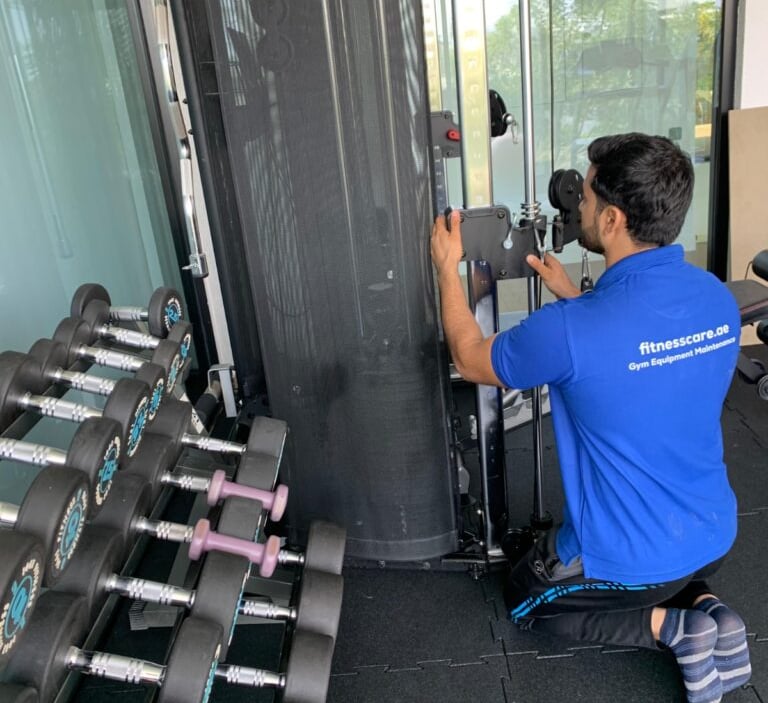 Professional technician performing gym equipment maintenance on a cable machine next to a dumbbell rack.