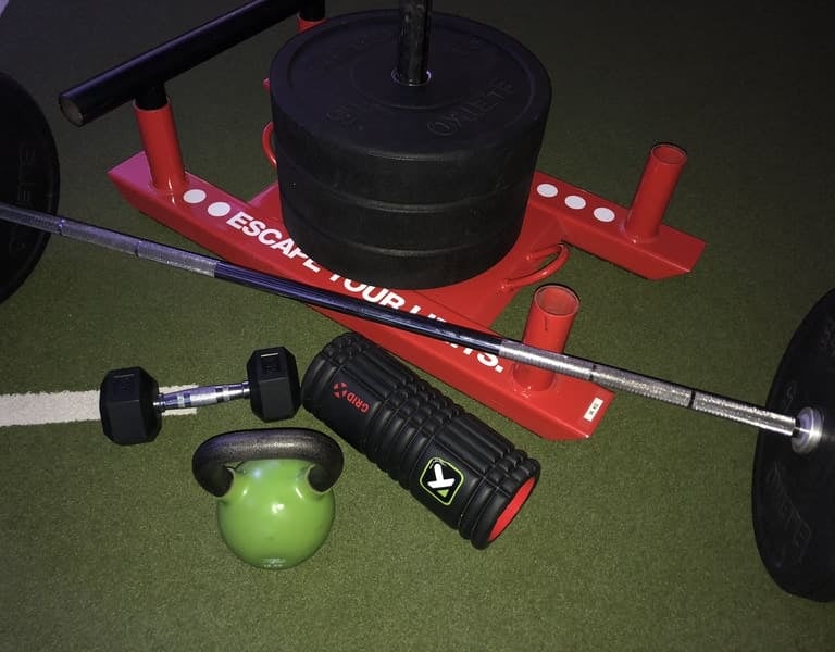 Fitnessgeräte auf Kunstrasen: roter Gewichtsschlitten, Langhantel, grüne Kettlebell und Faszienrolle