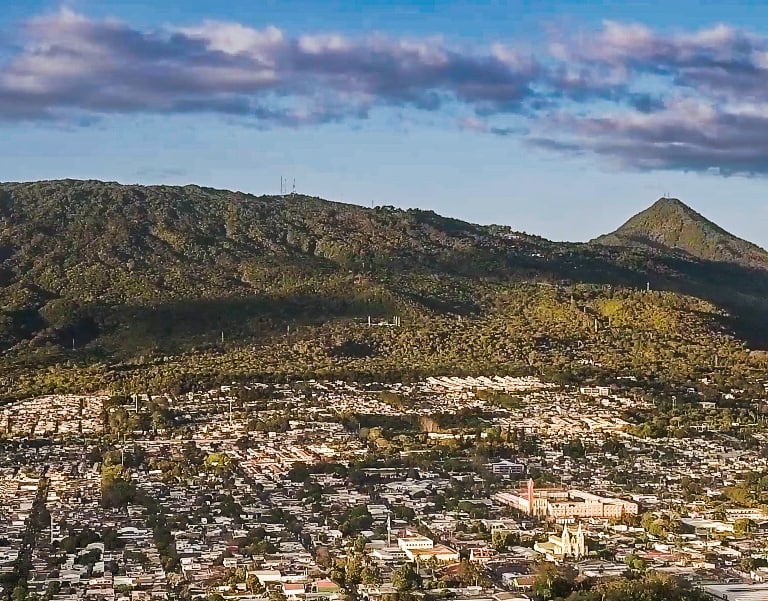Aerial view of Santa Tec;a nestled at the base of lush green volcanic mountains under a blue sky.