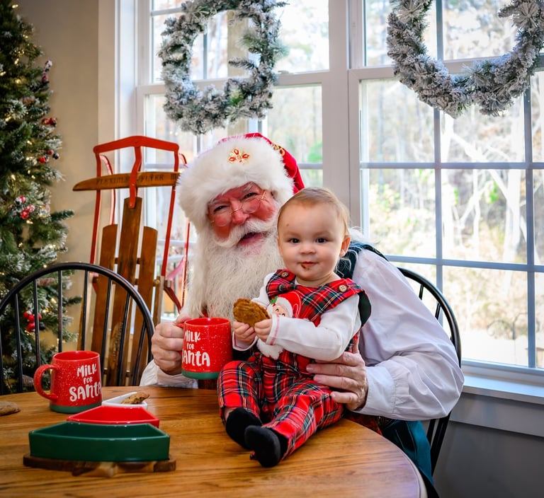 Santa Claus sitting at a wooden table with a baby in plaid overalls holding Christmas cookies and milk mugs.